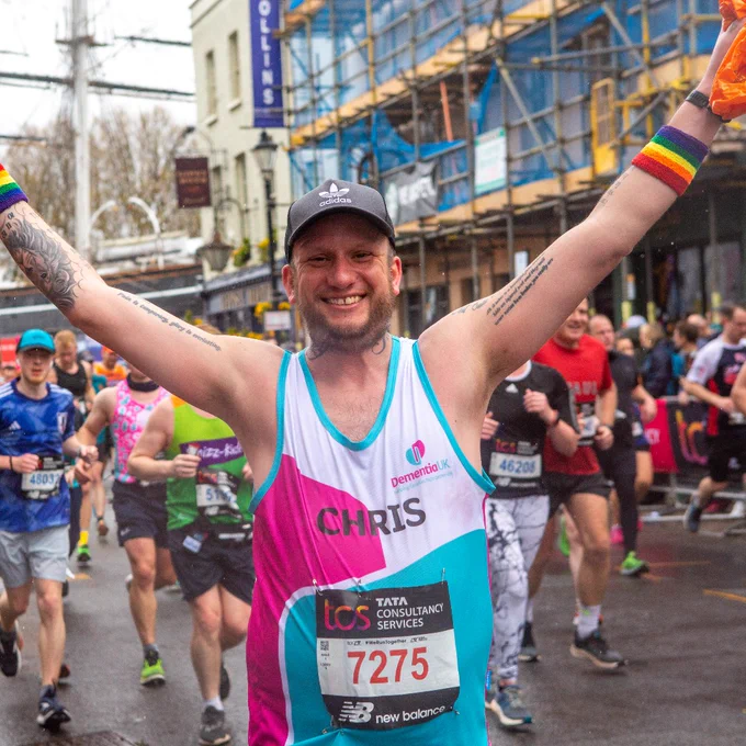 A group of runners on a road. A man wearing a Dementia UK running shirt is front and centre, smiling at the camera and raising his arms in the air in celebration. 