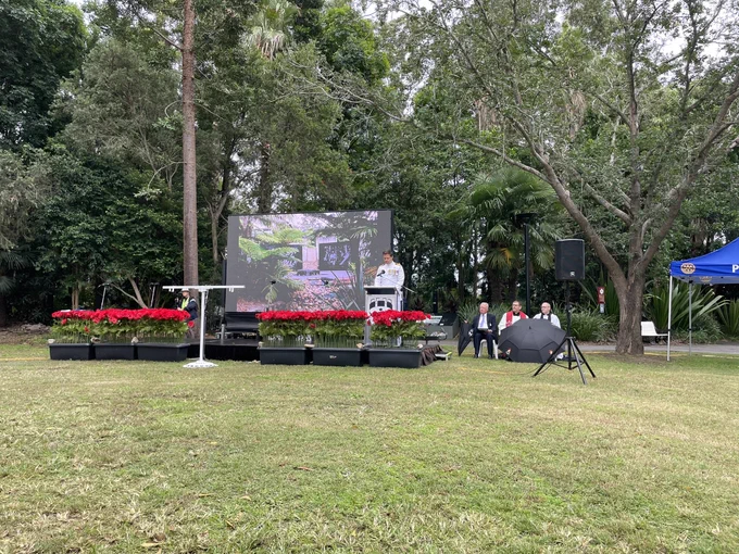 Captain Richard Brickacek of the Royal Australian Navy giving a speech in front of an outdoor screen showing the Kokoda memorial with a trees behind him and poppies in planters infront of him.