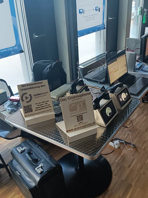 Ein Tisch, auf dem zwei hölzerne, gelaserte Werbetafeln für das Grannophone und den Makerspace stehen, sowie drei Grannophone in unterschiedlichen Bauzuständen. Unter dem Tisch ist ein lederner Pilotenkoffer zu sehen.