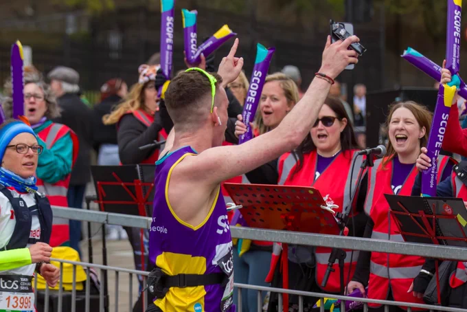A runner in a Scope running vest looks away from the camera at a group of singers behind a barrier at the London Marathon with his arms raised. The singers have microphones and are waving Scope inflatable batons. They are wearing pink hi-vis jackets and Scope t-shirts.