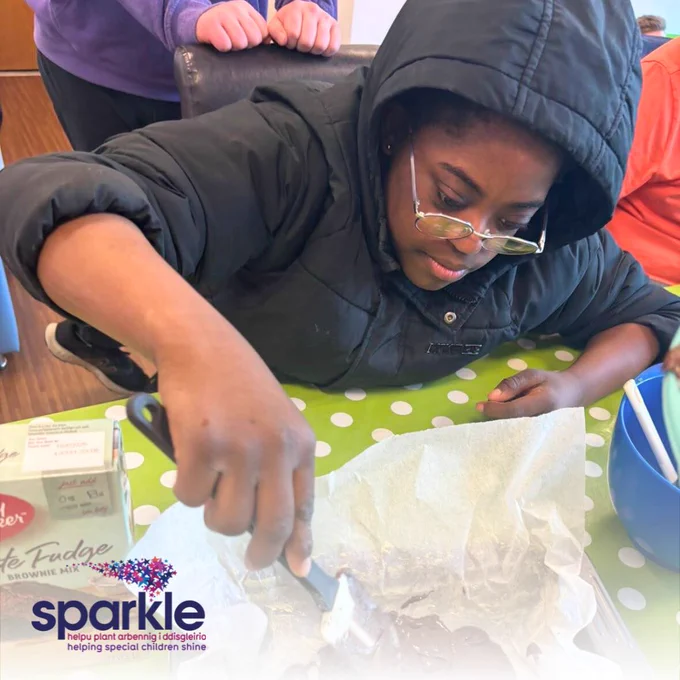 A young girl wearing a black coat with her hood up spreads brownie mix into a tray. 