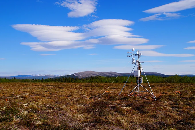A piece of equipment for measuring greenhouse gases, on a tripod in the Alaskan tundra, with mountains behind it
