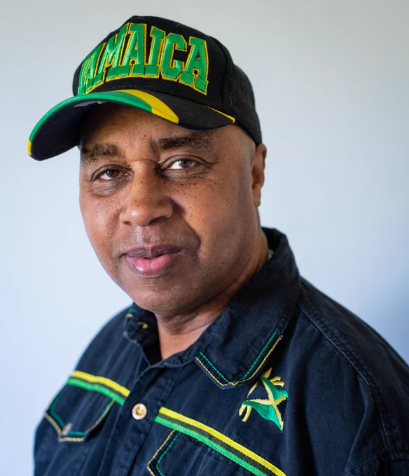 A portrait of Bob Clarke, founder of MAMA Youth project. He is wearing a black shirt with green, black and yellow stripes and embroidered detail and a Jamaican flag on the left chest. He is also wearing a black cap with the Jamaican flag across the peak and the word 'Jamaica' written in green with a yellow outline across the front of the cap.