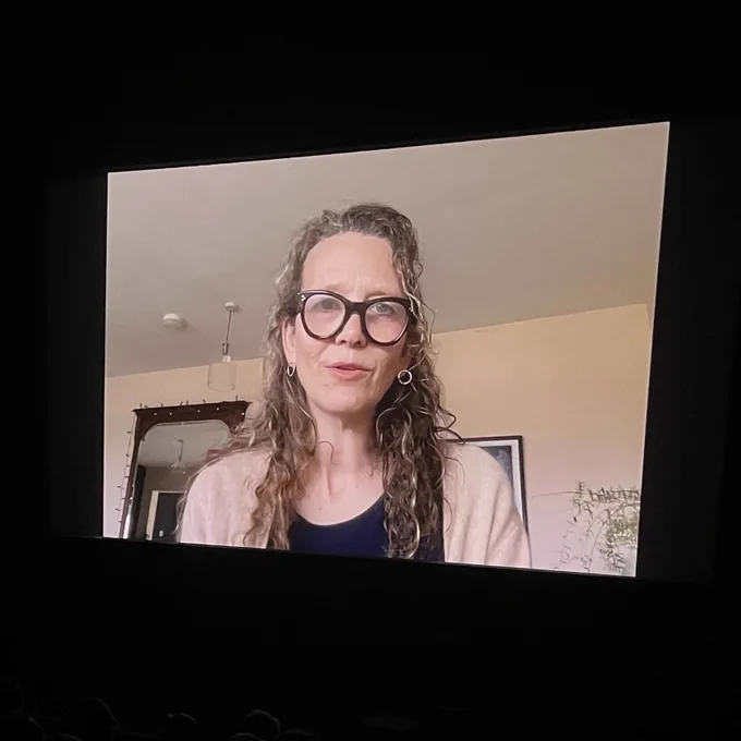 Film director Emma Davie speaking to camera, projected on a big screen.