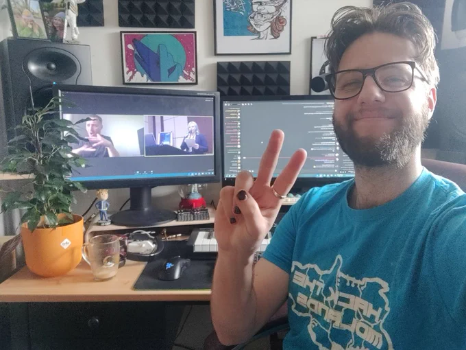 Luke takes a selfie at his desk. He is smiling and wearing a blue Hack The Midlands t shirt and has black painted nails. Behind him, a PC Monitor shows the Discord and Zoom call for the Game Accessibility conference. 