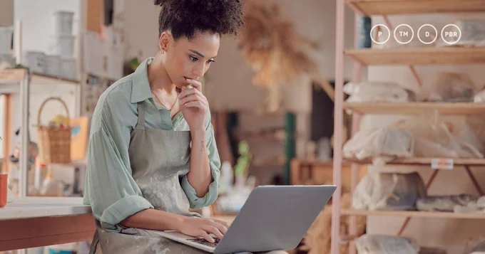 Women wearing a green shirt and apron sitting looking at a laptop. She is in an art studio.