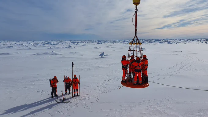 Seven researchers in bright orange overalls in an ice field in Antarctica. Three stand on the ground with spades and poles. Four stand on a platform lifted in the air from the ship (out of view). Find out more about the SEE researcher's Antarctica expedition: https://environment.leeds.ac.uk/faculty/news/article/5753/cold-unbalanced-and-feeling-seasick-all-in-the-name-of-science