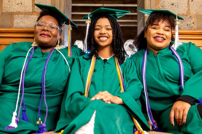 Three undergraduate students wearing their caps, gowns, and cords as they sit together for a photo.