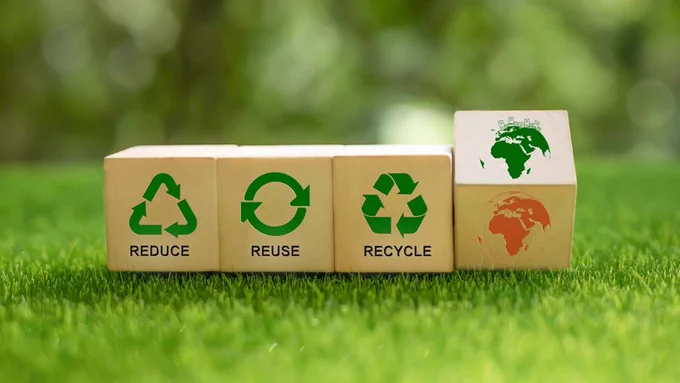 Four wooden blocks on grass with 'Reduce, Reuse, Recycle' symbols printed on them. A fourth block features a print of Earth turning from red to green.