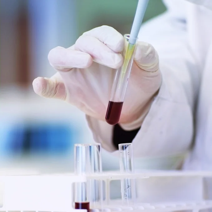 A gloved hand of a scientist holding a test tube and using a pipette to draw a sample of liquid.