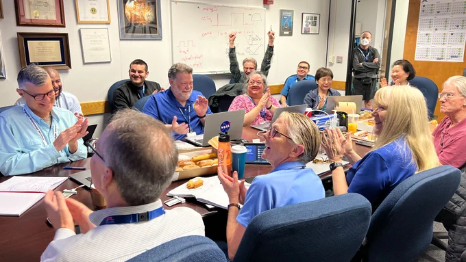 After receiving data about the health and status of Voyager 1 for the first time in five months, members of the Voyager flight team celebrate in a conference room at JPL. About 15 members of the team sit around a long conference room and are shown cheering and clapping upon receiving the good news.