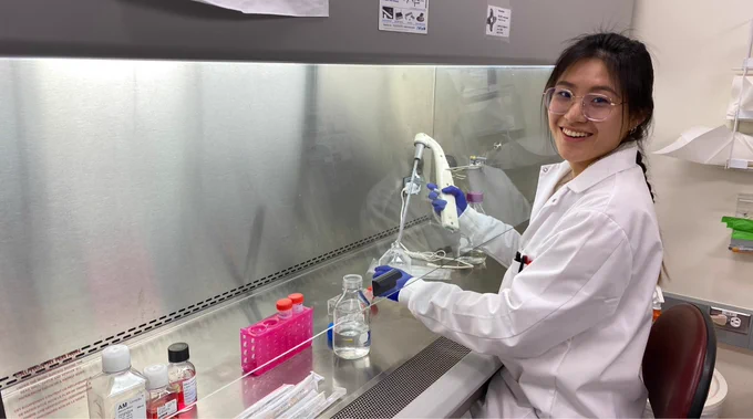 Foster lab graduate student Lucy Chi smiles to camera, holding a pipette to culture astrocyte cells in a biosafety cabinet.
