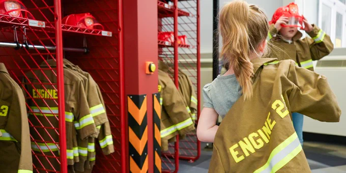 Photo of two children dressing up as fire fighters with jackets that read "Engine 23" and red fire fighter helmets. 