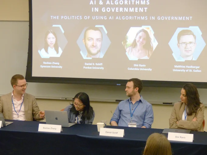 Participants in a conference panel speak while seated behind a table with paper name plates and a projection of their images above, with text: AI & Algorithms in Government, The Politics of Using AI Algorithms in Government, Baobao Zhang, Syracuse University; Saniel S. Schiff, Purdue University; Shir Raviv, Columbia University; Mathias Haslberger, University of St. Gallen