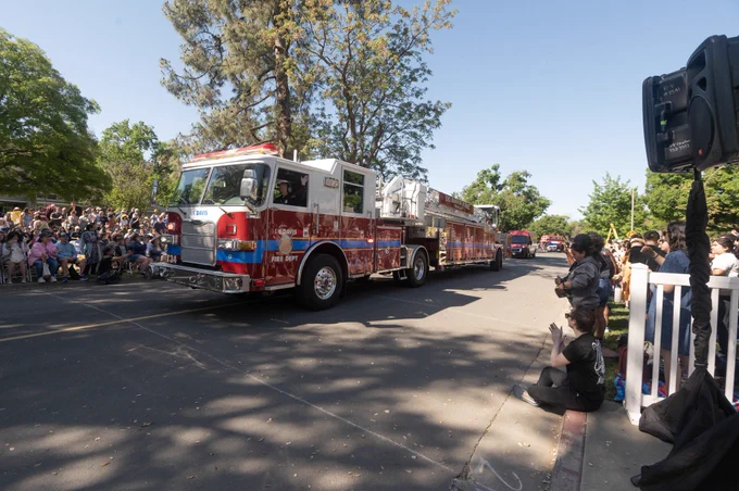 Truck 34 riding through the middle of a street with  the Health 34 van trailing. Pedestrians are on both sidewalks clapping, taking pictures, and waving. UCDFD personnel can be seen waving back at the crowd. Trees and natural elements are in the background.