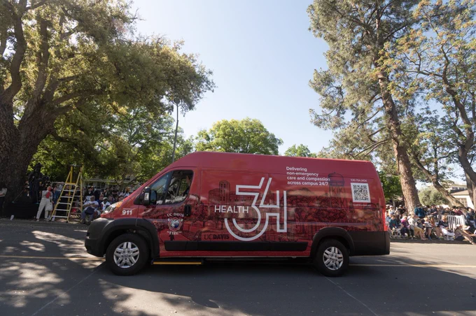 The Health 34 van in the middle of the street with pedestrians on the sidewalk admiring the van and upcoming parade participants. Trees and natural elements are in the background.