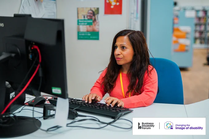 Professional who is short in stature works on a computer in a library. The Business Disability Forum Changing the image of disability logo is in the bottom right corner of the image.