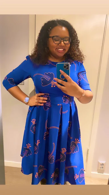 Dr. Angeline Dukes, a Black woman with curly hair and glasses, taking a selfie in a mirror before her keynote talk. She’s wearing a blue dress with pink patterns and looks very excited!