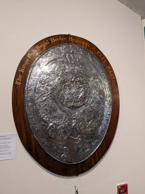 An elaborate metal shield depicting scenes from John Milton's Paradise Lost on a white wall in The Armitt museum. The metal looks silver, and is mounted on a wooden base, with gold gothic letters which read: The Kings Own Royal Border Regiment Fell Race