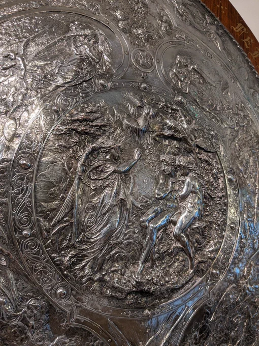 A close up of the shield's central panel, which shows the Archangel Raphael telling Adam and Eve of the war in heaven. Raphael stands on the lift in heavenly robes, wings on his back, telling the story to Adam and Eve, sat naked on a rock, opposite the angel, in the Garden of Eden