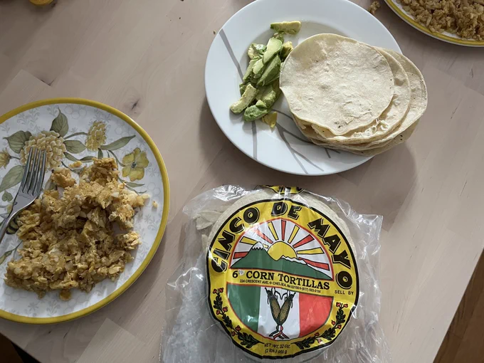 A bag of Cinco de Mayo brand corn tortillas sits on a table, beside plates of scrambled eggs, warmed tortillas, and sliced avocado.