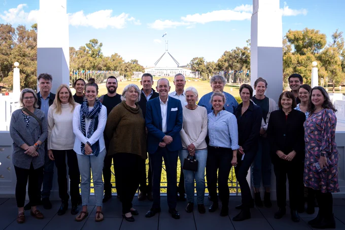 A group of 19 people stand shoulder to shoulder, facing the camera, with Parliament House in the background. 