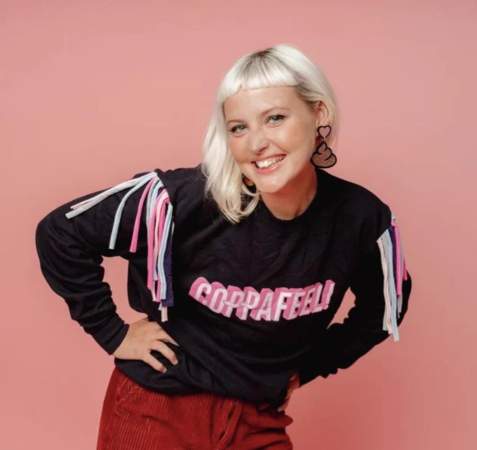 Photo shows Kris Hallenga smiling at the camera with her hands on her hips, wearing a CoppaFeel! jumper and poo-emoji earings.