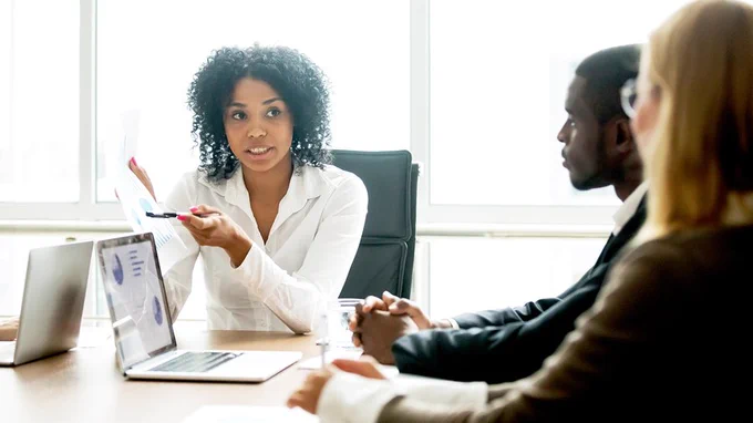 Two people sitting at an office table with a laptop in front of them listing to the woman at the end of the table.