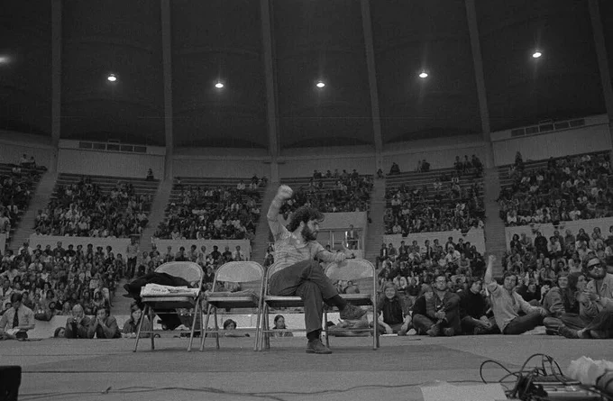 Black and white photo of Jerry Rubin on stage with his fist in the air before a crowd of UVA students gathered at University Hall. May 6, 1970 (RG-5/7/2.762)