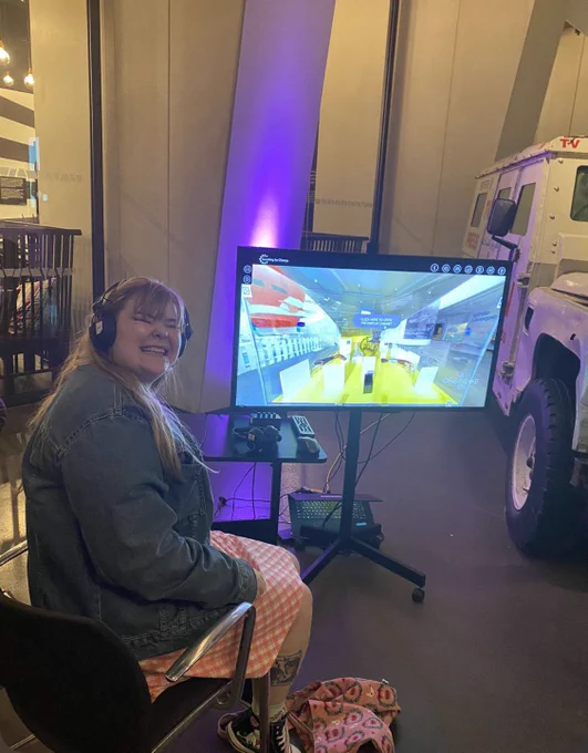 Iris smiling as she watches a 360 tour of her temporary exhibition at Museum of Liverpool on a large screen. She has headphones on to listen to an audio description of the display case, which includes different pieces of assistive technology.