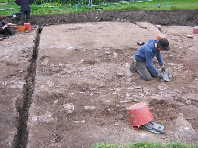 A student is kneeling and using a trowel to uncover a spread of roof slates.