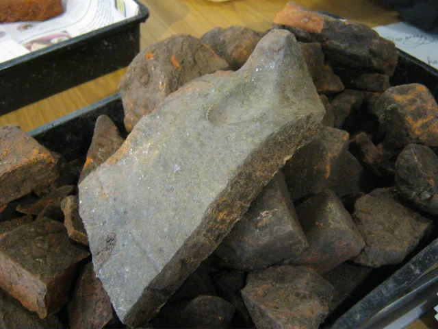 A tray filled with broken tiles, on top is part of a glazed ridge tile with an upstanding crest on its apex, one side of the ridge is missing.
