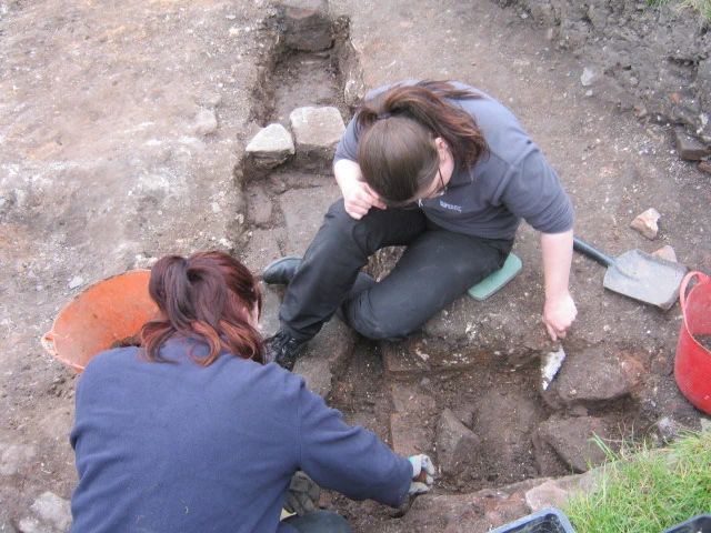 Two students are excavating a narrow trench filled with stone blocks, the foundations for a wall.