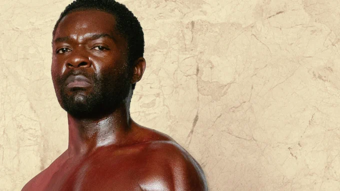 A portrait of a shirtless man with a muscular build, looking seriously at the camera, against a textured beige background.