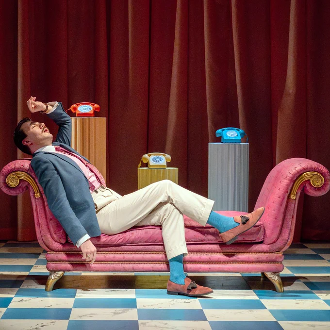 Actor Alex Phelps on the stage of Tilted Wig's production of The School for Scandal. 
Phelps is wearing a beige suit with a grey coat and a pink tie. He is lying down on a pink sofa. He is looking distressed, with one hand raised close to his forehead.
In the background are three vintage colourful telephones placed on a podium each of different heights. 