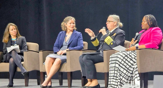 Four professionals sitting on stage engaged in a panel discussion, with one woman gesturing while speaking and three women listening attentively.
