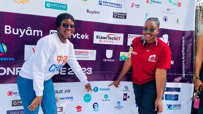 The Two ladies co-founders of Traleor pointing at the Traleor logo on a banner of logos during the Silicon Mountain Conference 2023
