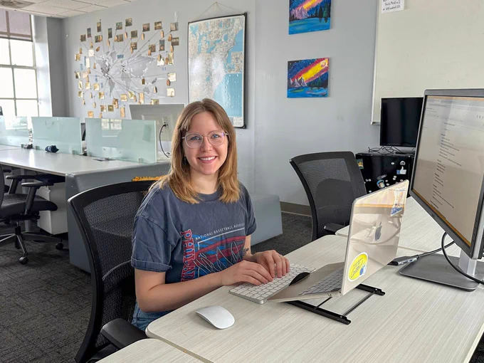 Woman with glasses smiling at her desk in a bright office, working on a computer.