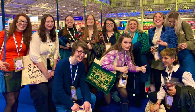 Group photo of 8 people standing in a line & 3 people crouching or semi-sitting in front. Various attire, all smiling. (Industrial-looking backdrop is London Book Fair.)