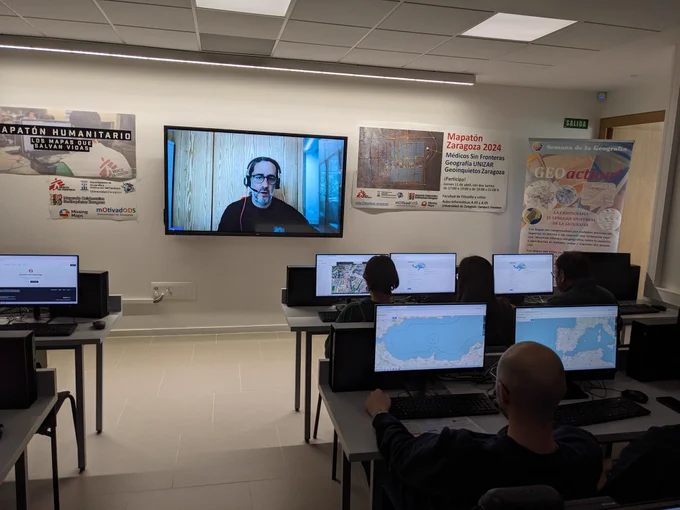 Sala de ordenadores con los participantes del mapaton y al fondo pantalla con la cara de Paco Arévalo de Médicos sin Fronteras presentando la actividad
