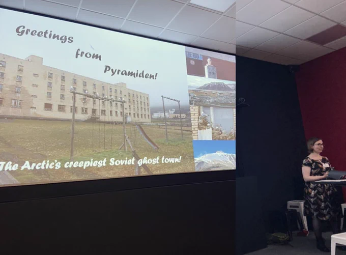 Fionnuala McCully standing by a slide with a concrete building reading “greetings from pyramiden! The Arctic’s creepiest soviet ghost town” including a photo of a bust of lenin