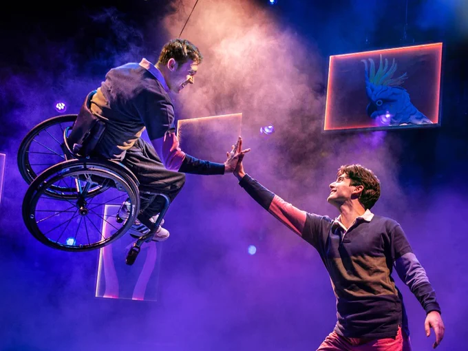 A man in a wheelchair flies in the sky and touches palm to palm with a man stood on the floor. In a theatre with stage smoke and blue lighting.