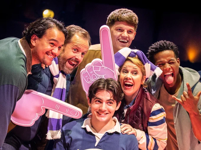 A group of people pose for a photo with giant pink foam fingers and sport scarfs.