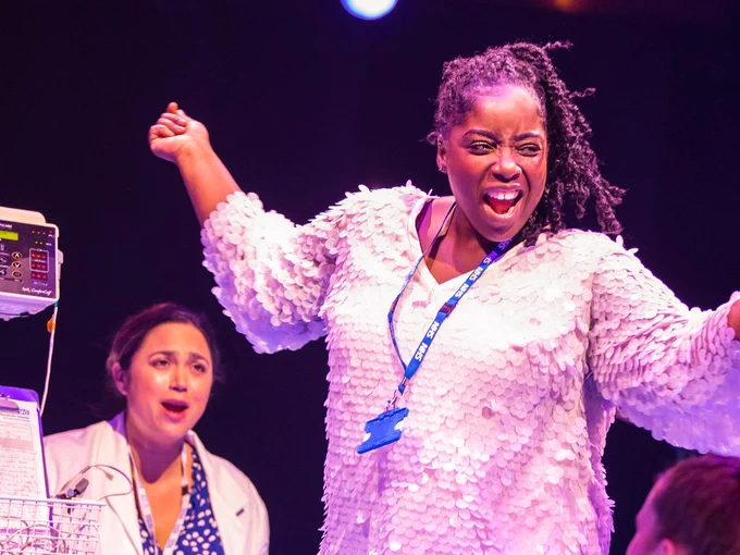 A woman in a white top and nhs lanyard dances. Behind her is a woman in a blue dress and white doctors coat.
