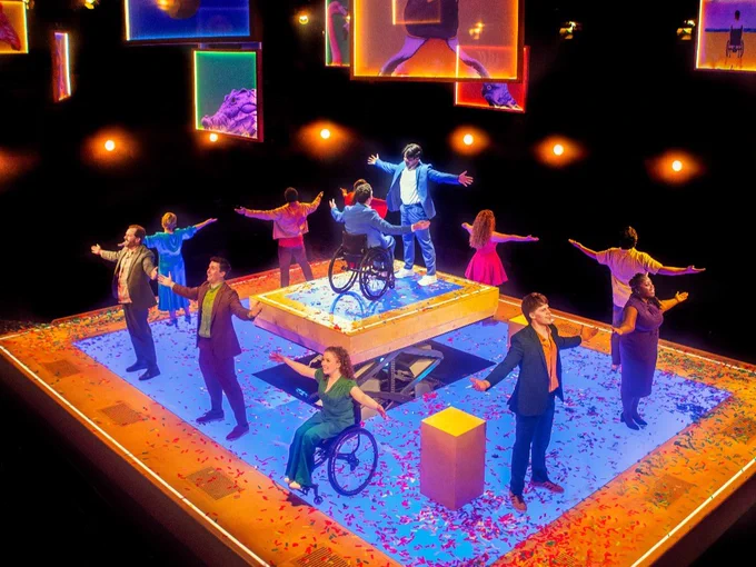 A theatre stage set. People stand around it with their arms out signing. From the ceiling hangs perspex slides in different colours.
