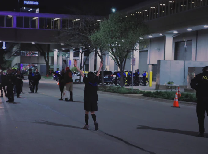 a de-arrested labor activist, walking back to their friends after being released by the cops. they are holding their hands above their arms, and giving 2 peace signs.
