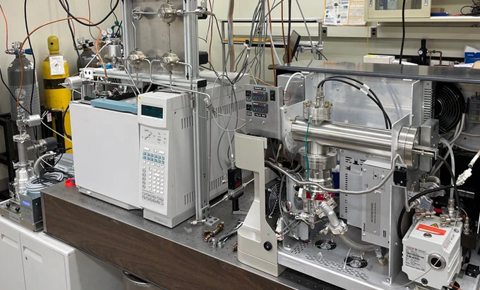 Two square machines sit on a lab table, one sheathed in white plastic and the other metal. Both have complex attachments, wires and connectors.