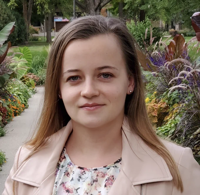 A woman in a floral shirt and pink jacket smiles serenely at the camera. She is standing in a garden.
