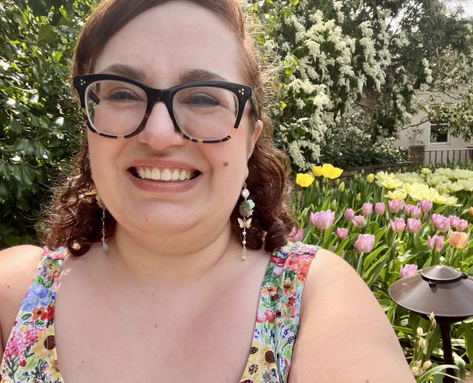 Daniele is a short, fat white woman with curly auburn hair and black glasses. She grins, wearing a rainbow-floral dress and standing in front of a tulip garden. End alt text.