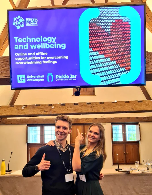 Simon Fairbanks and Celine Kuysters with a screen saying, "EFMD Technology and wellbeing, online and offline opportunities for overcoming feelings," beside a digital heart.